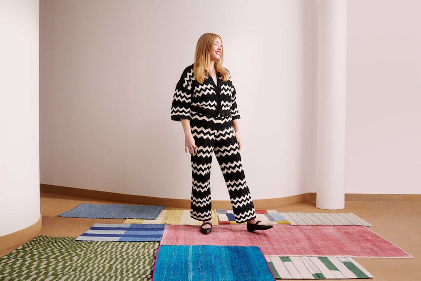 A picture of Emma on a photoshoot in a stripy outfit stood on some colourful rugs.