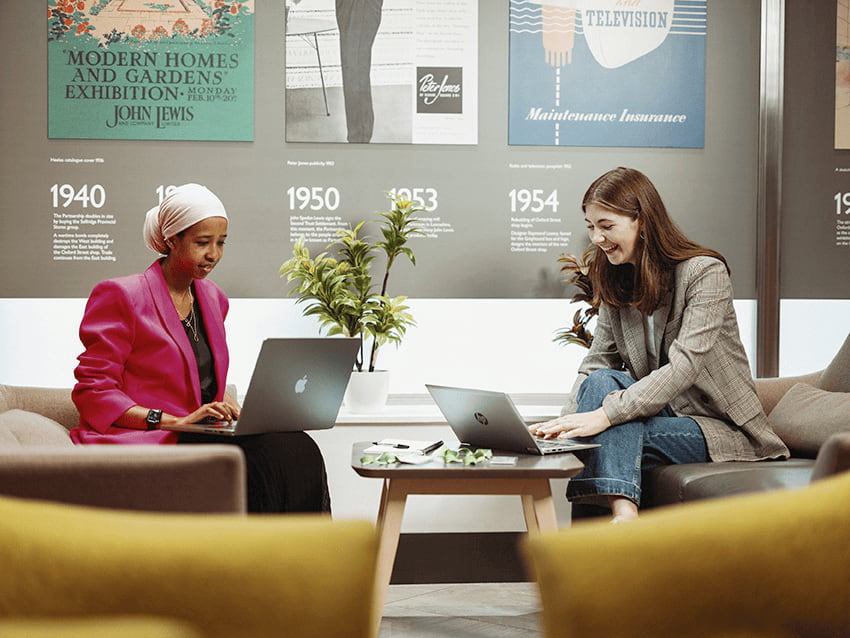 Two female John Lewis employees sat apart from each other with laptops smiling