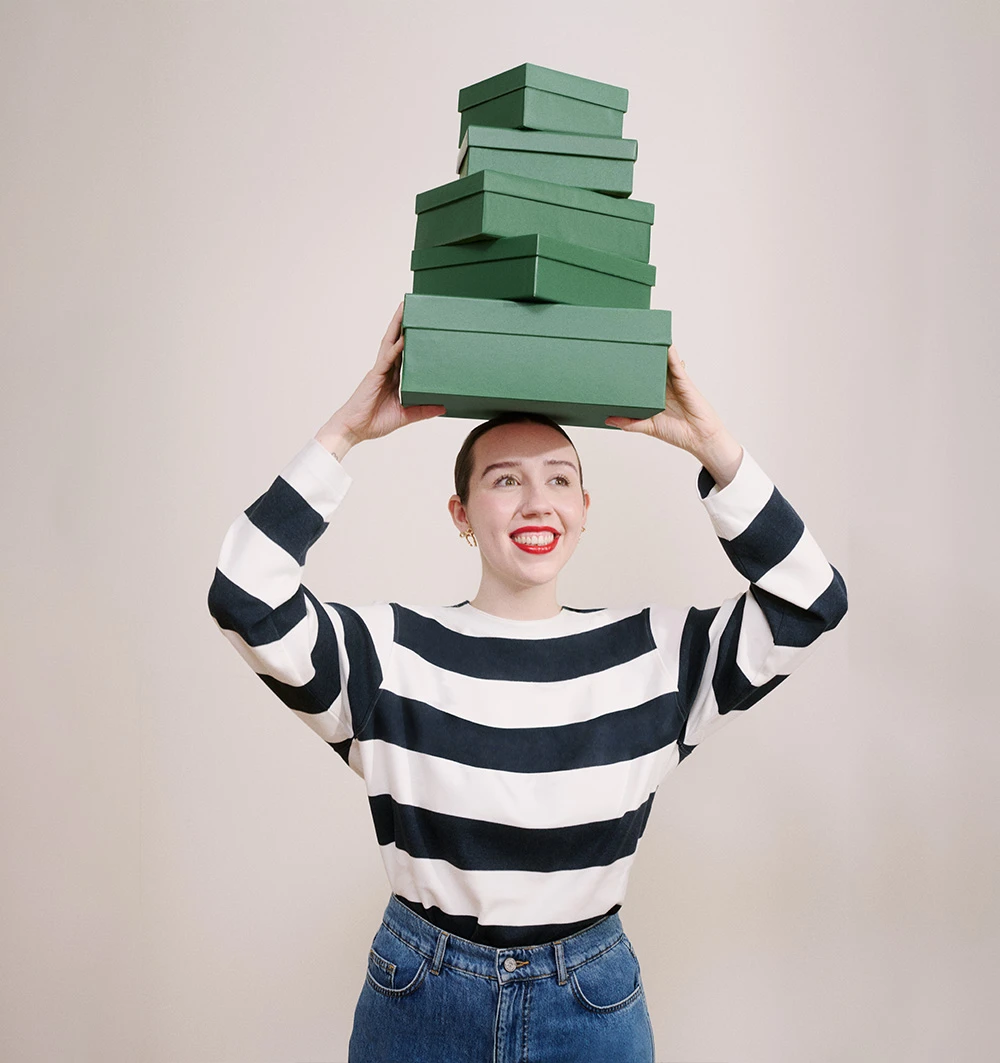 Vanessa in a striped jumper holding a stack of green boxes above her head.