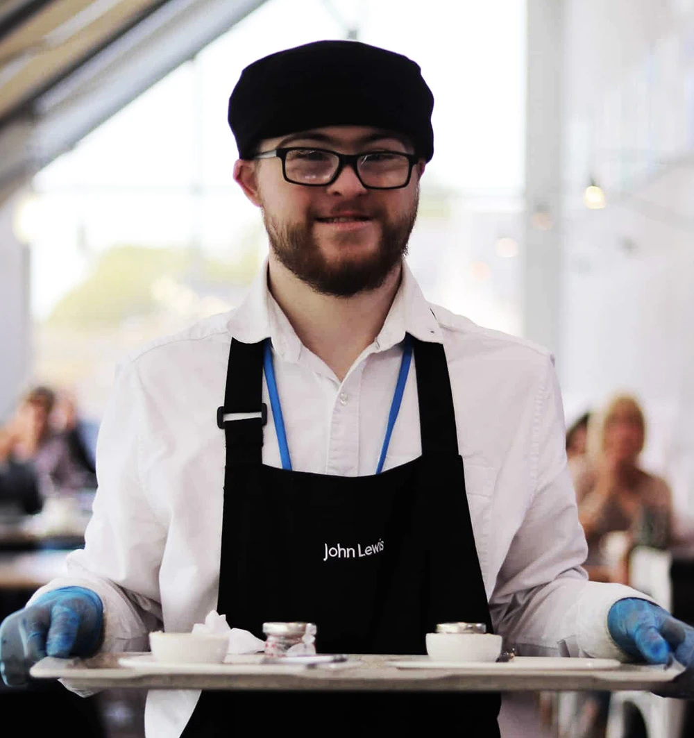 Catering assistant carrying a tray of food across a busy cafe