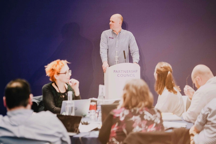 Man stood up in front of a group of people introducing Partnership Council with a purple background.