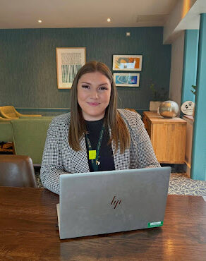 Anna sits in a lounge with her silver laptop on a table in front of her. She has straight, brunette hair, and wears a smart grey blazer with a black top.