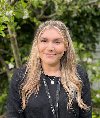 Anna is smiling to the camera, she is outdoors in front of a backdrop of greenery. She is wearing a black, long sleeved top, with a necklace and a John Lewis lanyard.