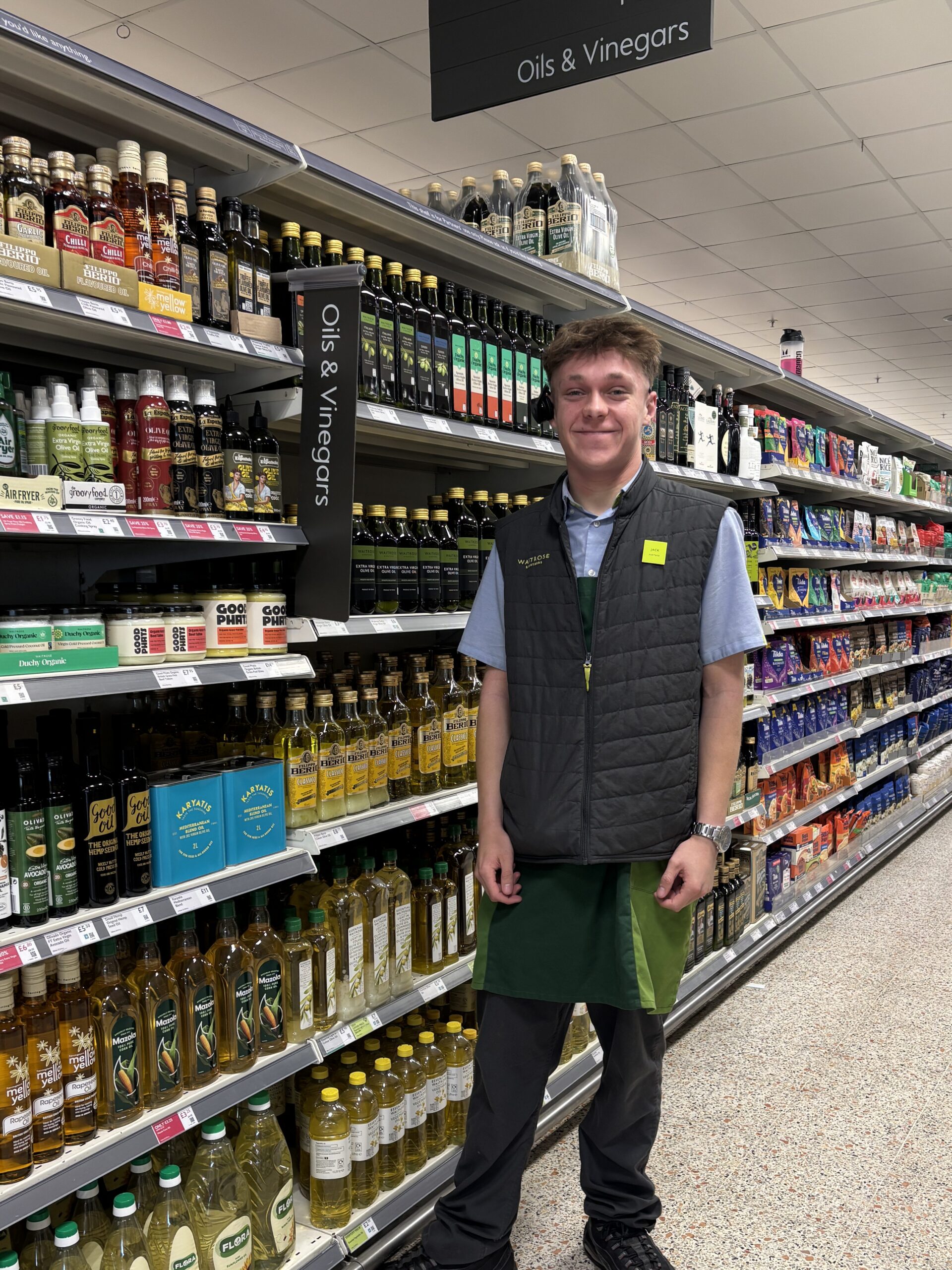 Young man with short blonde hair stands in a Waitrose shop. He is smiling and wears a Waitrose uniform.