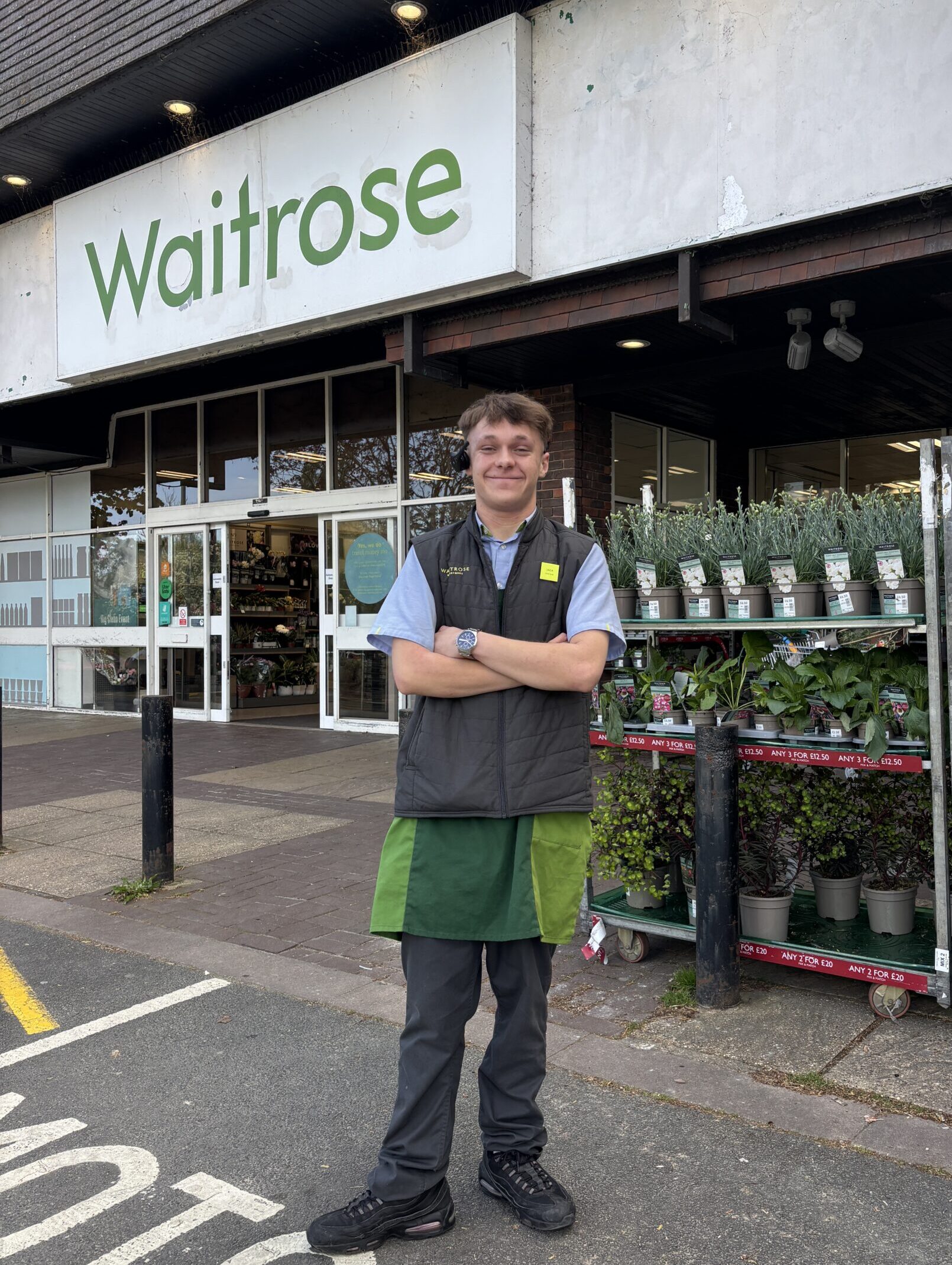 A young man with short blonde hair stands in front of a Waitrose shop. He is smiling and his arms are folded, he wears a blue shirt, grey gilet and black trousers. Behind his are green plants for sale at the shop.