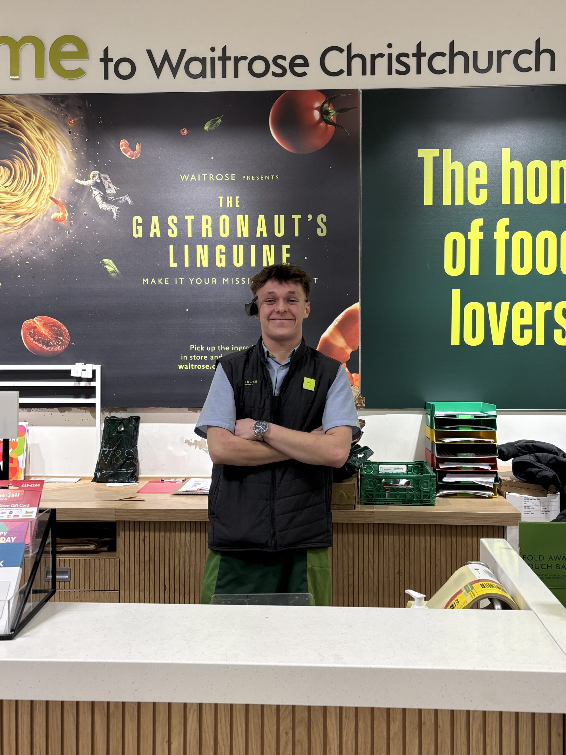 Image of young man in a Waitrose uniform, standing at a Welcome Desk in a Waitrose shop. He is smiling with his arms folded.