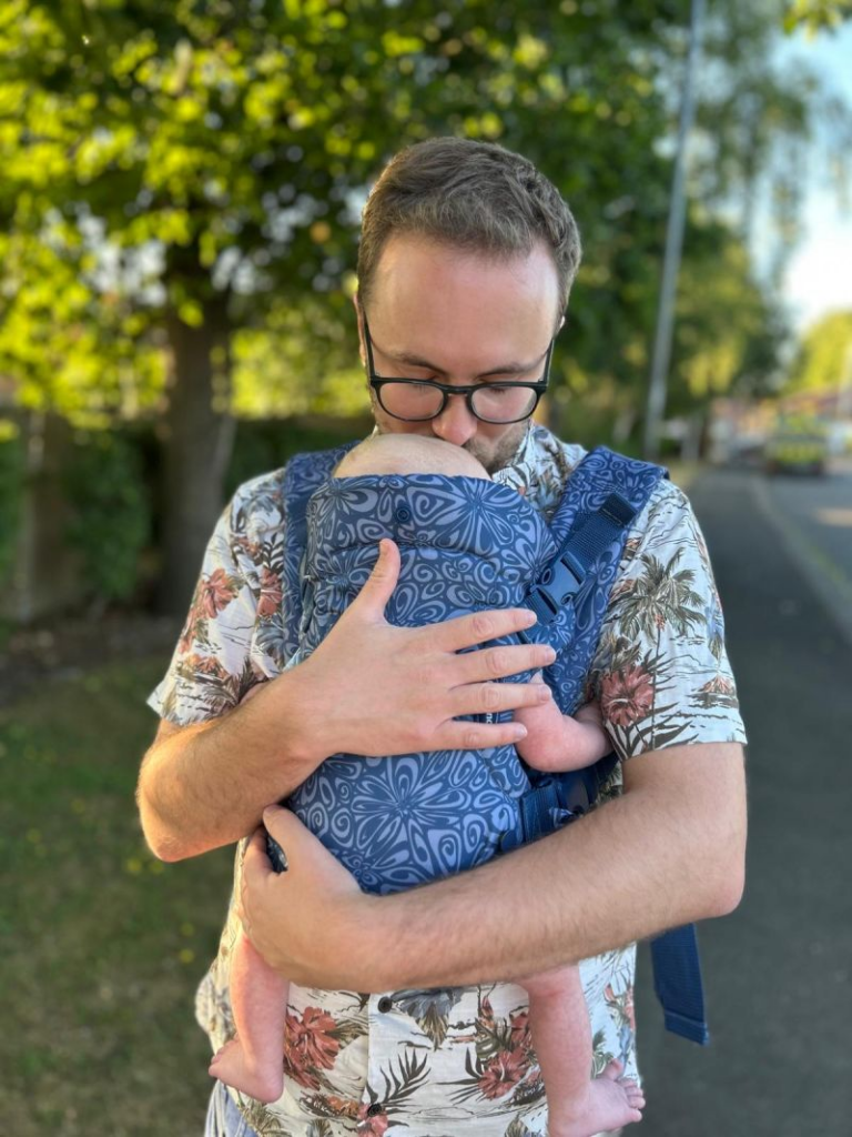 A white man with blonde hair holds his baby in a blue carrier. The man wears a floral shirt and dark-rimmed glasses. The baby is held in a blue, patterned baby carrier, which the man wears on his front. They are outside and walking along a road, with green trees and foliage to the left of the image, and the pavement to the right hand side.