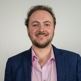 A white man is smiling to the camera. He wears a dark blue blazer and a pink shirt. He has a beard and moustache, and short hair.