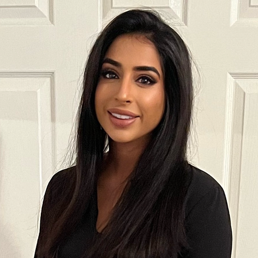 Young, British-born South Asian Muslim woman smiles to the camera. She is wearing a black top, has long, straight dark hair, and is stood in front of a white door.
