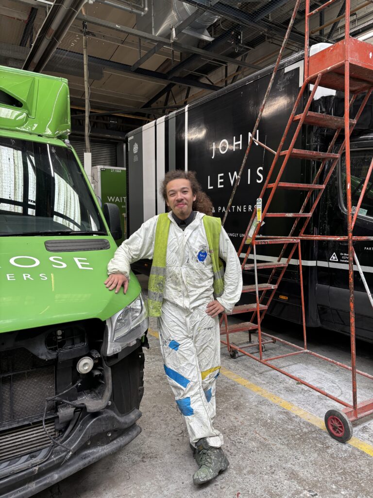A smiling worker in white protective clothing and high-visibility vest stands between a green Waitrose delivery van and a black John Lewis truck inside a large garage.