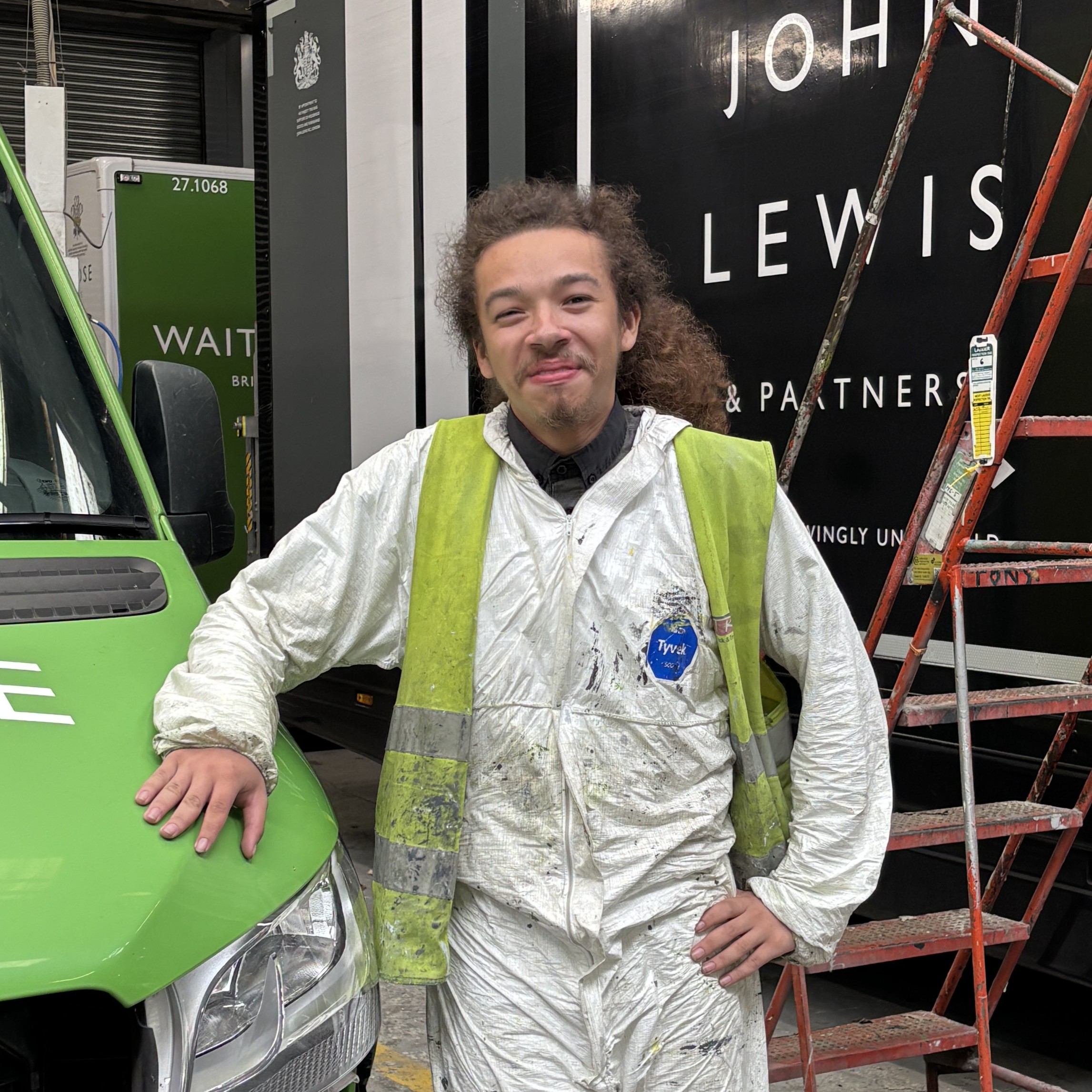 A smiling worker in a white hazmat suit and high-visibility vest stands between a green Waitrose delivery van and a black John Lewis truck inside a workshop.
