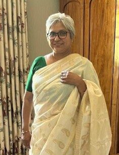 A lady with short, grey hair and of Hindu and Malaysian heritage, looks to the camera and smiles. She is wearing dark-framed glassed, a green short sleeved top and a white and gold traditional saree.